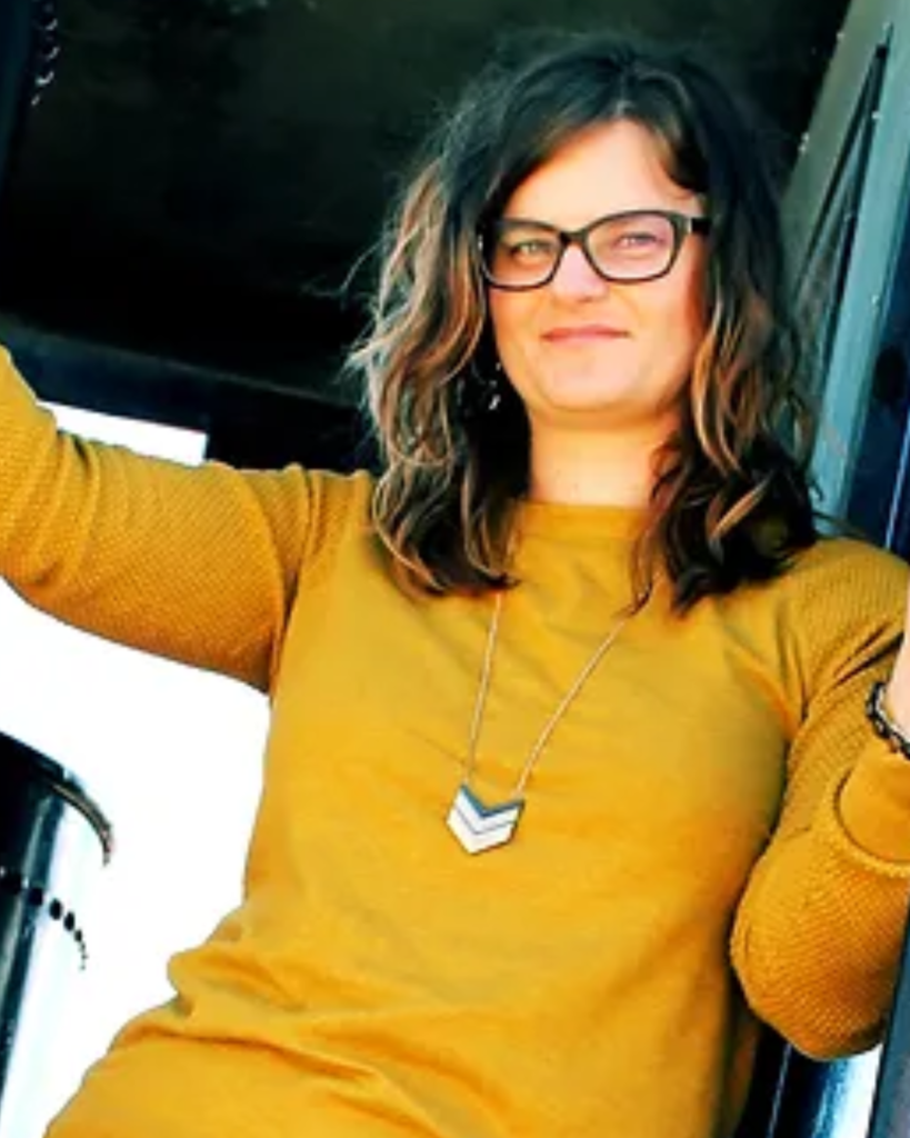 Photo of young white woman with glasses and curly hair wearing a yellow sweater.